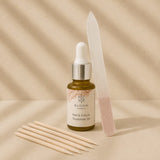 Bloom Nail & Cuticle Treatment Oil in a small amber dropper bottle, styled with wooden cuticle sticks, a translucent plastic nail file, and pink nail polish on a warm beige background with soft shadow overlays.