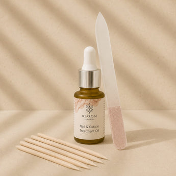 Bloom Nail & Cuticle Treatment Oil in a small amber dropper bottle, styled with wooden cuticle sticks, a translucent plastic nail file, and pink nail polish on a warm beige background with soft shadow overlays.
