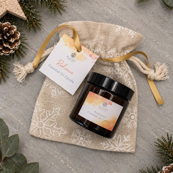 A small amber glass essential oil candle jar with a black lid sits on a beige burlap drawstring pouch with white snowflake print. A white hanging tag reads 'Relapse Essential Oil Candle' and shows the Melrose & Co brand.