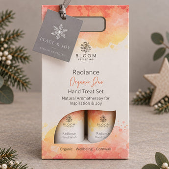 Bloom Remedies Radiance Organic Duo Hand Treat Set in a watercolor orange and yellow gift box with a hanging grey tag, styled with pine branches and a star ornament on a neutral background.