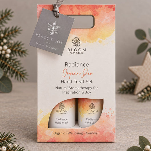 Bloom Remedies Radiance Organic Duo Hand Treat Set in a watercolor orange and yellow gift box with a hanging grey tag, styled with pine branches and a star ornament on a neutral background.