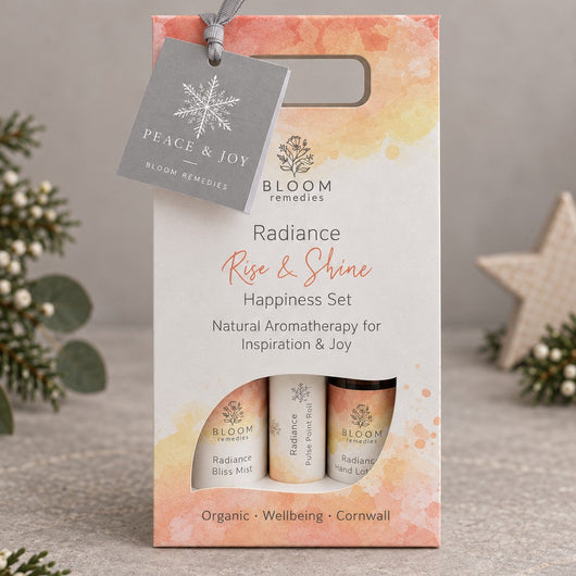 Bloom Fragrances Radiance Happiness Set in a watercolor orange and cream gift box with a hanging grey tag, displayed on a neutral grey surface with decorative pine branches and a star ornament.