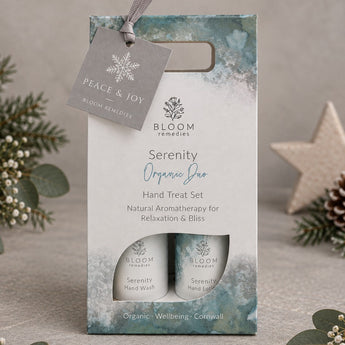 Bloom Remedies Serenity Organic Doo Hand Treat Set in a decorative gift box with two small cylindrical bottles visible through a window, plus a hanging tag reading Peace & Joy, styled on a neutral surface with greenery and a star ornament.